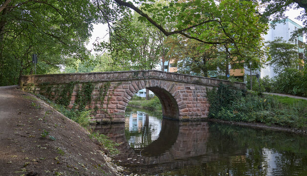 An old stone arch bridge crossing a calm canal surrounded by lush green trees in a park setting