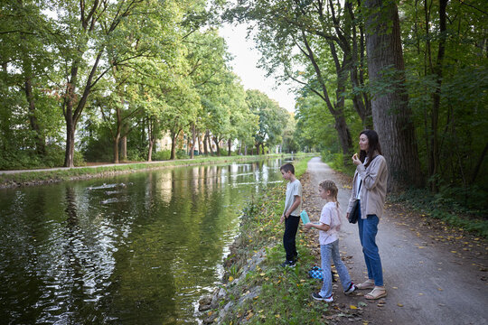 A mother and her two children standing by a peaceful canal in a lush green forest park setting