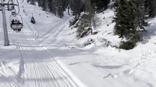 Riding a chairlift moving upwards over deep snow covered mountain slopes and ski tracks with evergreen pine trees under bright sunlight on a sunny winter day.