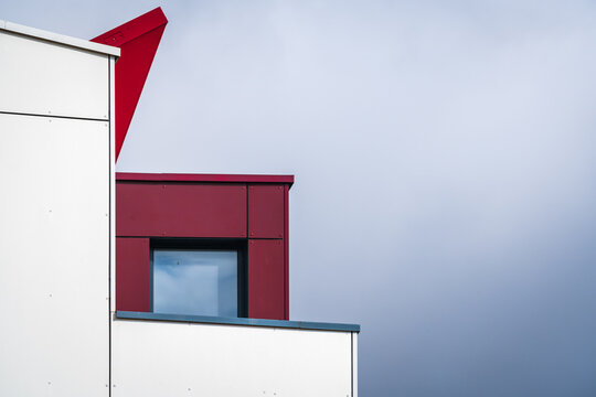 Modern minimalist rooftop geometry of stacked white building facades with flat parapets, small recessed window, and bold red angled accent under overcast sky in urban exterior composition