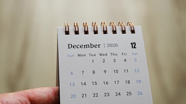 Hand holding desk calendar showing December 2026 page with dates and weekday columns on neutral background