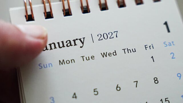 Finger tracing dates on desk calendar page showing January 2027 with weekday columns in close up
