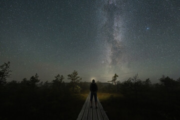 Fototapeta premium Man standing on wooden boardwalk in dark bog facing starry sky with Milky Way galaxy in Estonia nature reserve during clear night.