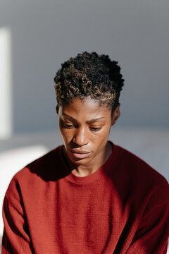 Pensive young black person looking down in warm directional sunlight wearing a red knit sweater for introspective portrait with soft shadows