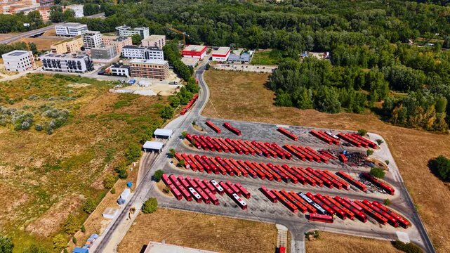 Large bus station with numerous vehicles standing outdoors. Green forest, multi-storied buildings and waste water reservoirs around the depot.