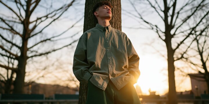 Contemplative young man in oversized shirt leaning against tree at sunset with soft backlight for outdoor portrait background and moody natural tones