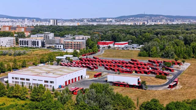Lots of red city buses stand in rows outdoors near the depot building. Lush green forest, a cityscape and green hills in the backdrop.