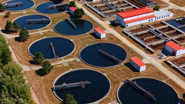 Flight over several huge round sewage reservoirs among few trees. Territory of modern waste water treatment plant at daytime.