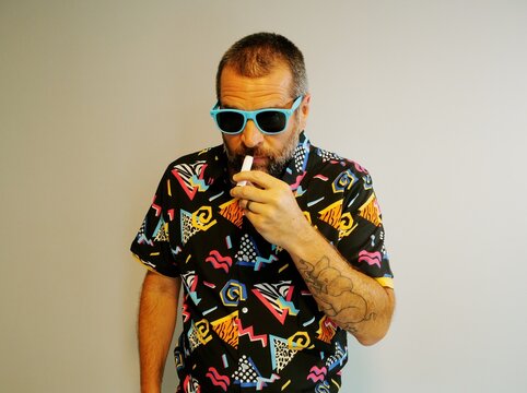 Middle aged man in 1980s graphic pattern shirt neon sunglasses and fanny pack posing humorously trying to look cool on neutral studio background