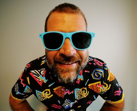 Middle aged man in 1980s graphic pattern shirt neon sunglasses and fanny pack posing humorously trying to look cool on neutral studio background