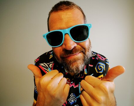 Middle aged man in 1980s graphic pattern shirt neon sunglasses and fanny pack posing humorously trying to look cool on neutral studio background
