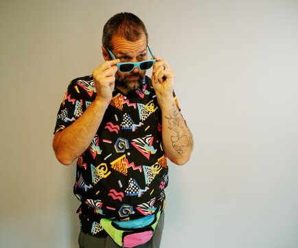 Middle aged man in 1980s graphic pattern shirt neon sunglasses and fanny pack posing humorously trying to look cool on neutral studio background
