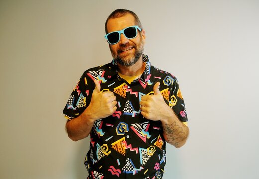 Middle aged man in 1980s graphic pattern shirt neon sunglasses and fanny pack posing humorously trying to look cool on neutral studio background