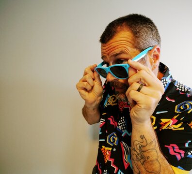 Middle aged man in 1980s graphic pattern shirt neon sunglasses and fanny pack posing humorously trying to look cool on neutral studio background