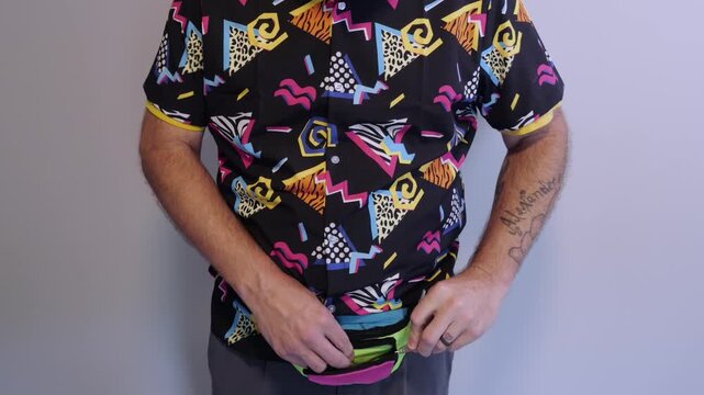 Middle aged man in 1980s graphic pattern shirt neon sunglasses and fanny pack posing humorously trying to look cool on neutral studio background