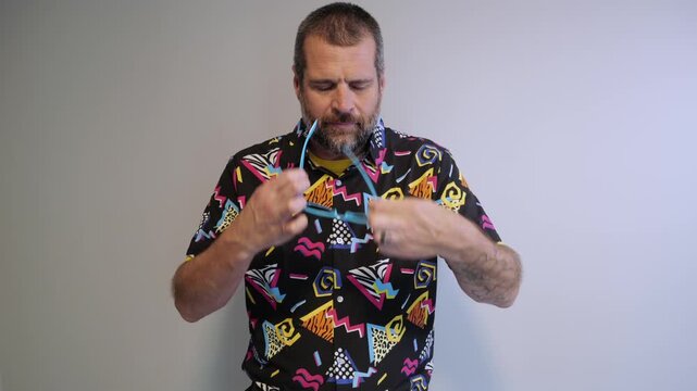 Middle aged man in 1980s graphic pattern shirt neon sunglasses and fanny pack posing humorously trying to look cool on neutral studio background