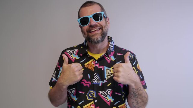 Middle aged man in 1980s graphic pattern shirt neon sunglasses and fanny pack posing humorously trying to look cool on neutral studio background