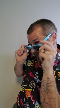 Middle aged man in 1980s graphic pattern shirt neon sunglasses and fanny pack posing humorously trying to look cool on neutral studio background