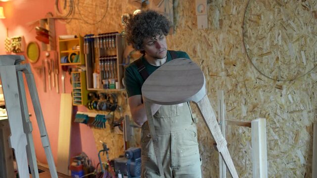 Private production of wood products, focused man carpenter examines semi-finished chair. Craftsman holding stool and precisely checking quality of wooden elements, crafter proud of new item. 