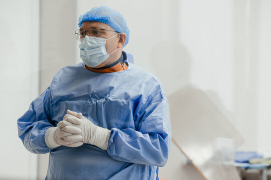 Senior surgeon in sterile gown, face mask and gloves standing with clasped hands in operating room, ready for procedure, copy space. Mature male doctor waiting before cardiology procedure in hospital