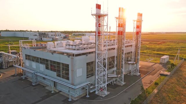 Top-down aerial panorama of modern industrial plant featuring three tall red and white smokestacks, steel towers, rooftop pipes, ladders, and walkways glowing in sunset light over facility yards
