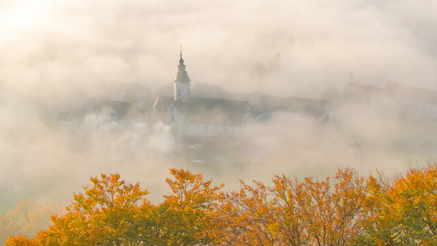 AERIAL, CLOSE UP: Historic Sticna Abbey appears almost dreamlike through dense morning mist, with its distinctive steeple rising above pale fog and warm autumn foliage in peaceful Dolenjska, Slovenia.