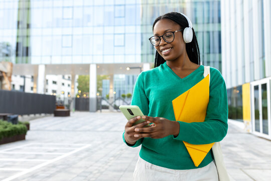 Smiling black student listening music using smartphone and headphones outdoors