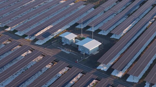 Aerial view of vast solar farm rows. White buildings sit center, surrounded by panels. Sunlight glints off each solar unit clearly. No people seen, no animals present. Ideal for green energy