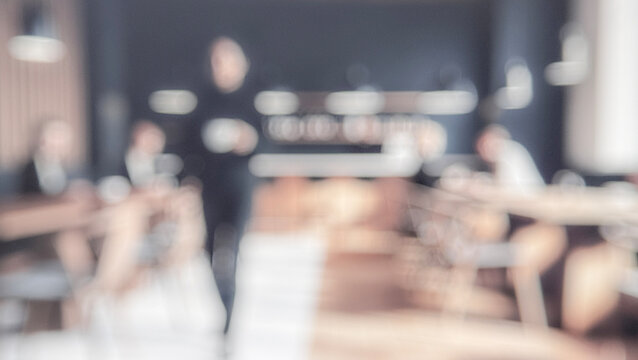 A blurred view of a modern office or cafe, featuring a person carrying a drink in the foreground, with several individuals seated at tables in the background.