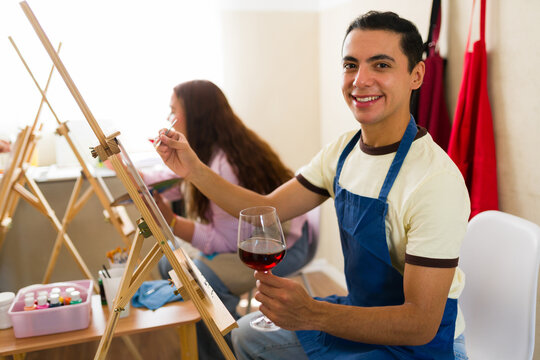 Portrait of a man enjoying an adult painting class, holding a glass of wine while creating art with friends