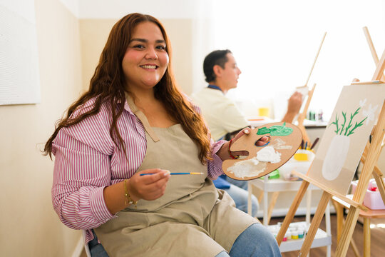 Happy plus size woman learning to paint in an adult art class, holding a palette and brush, enjoying creativity and hobby
