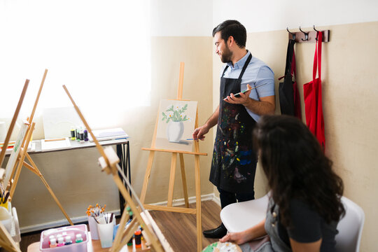 Male art instructor teaching painting techniques to a female student in an adult art class workshop studio