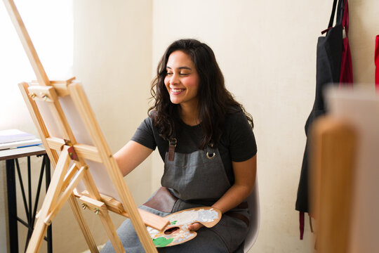 Smiling young woman painting on a canvas, holding a palette with paint colors, enjoying a creative art class