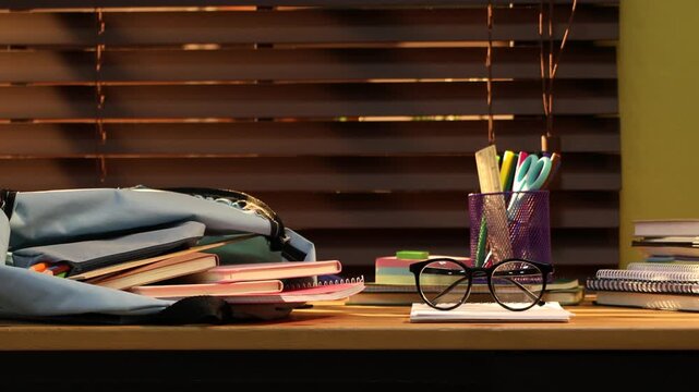 Stylish backpack with different stationery, glasses and apple on wooden desk indoors, camera moving left
