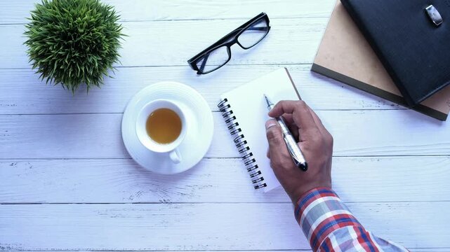Top-down view of a male hand, writing on a small notepad with a pen, next to a cup of tea, eyeglasses, and books on a white wooden table