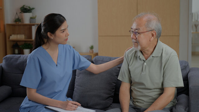 Caring Asian female nurse in blue uniform gently touching senior man shoulder with empathy, offering support during home healthcare checkup visit in living room.