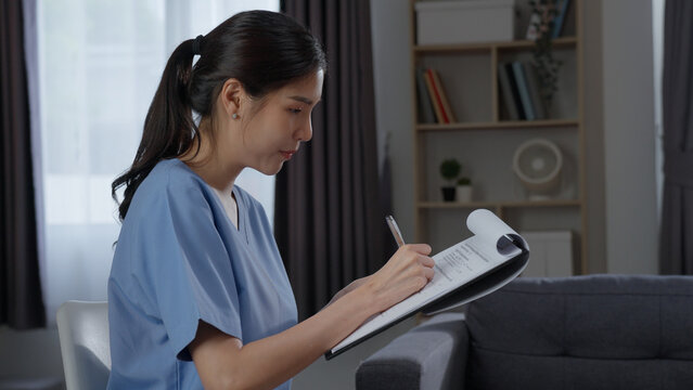 Side profile view of focused Asian female nurse sitting and writing patient medical report on paper clipboard with silver pen during healthcare home consultation.