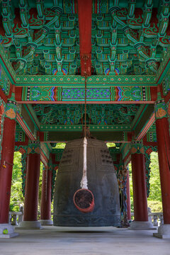 Large traditional bronze bell hanging beneath intricate colorful dancheong wooden ceiling in Buddhist temple bell pavilion in South Korea.