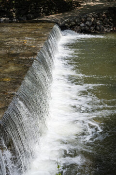 Clear fresh stream water cascading over stone weir creating white foam in peaceful outdoor nature landscape.