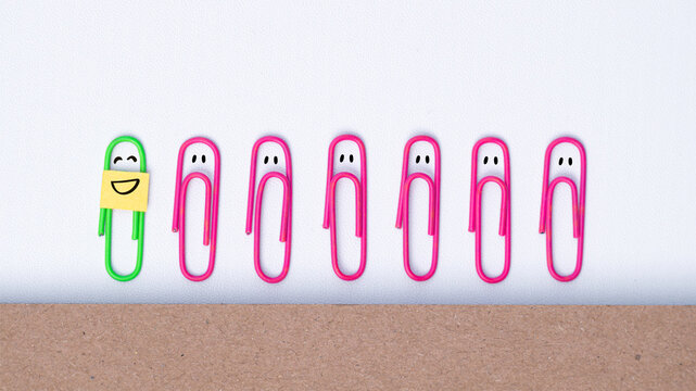 Cheerful smiling paper clip standing among several sad paper clip characters, creating a conceptual message about happiness, optimism, resilience, positivity, and finding joy despite difficult emotion
