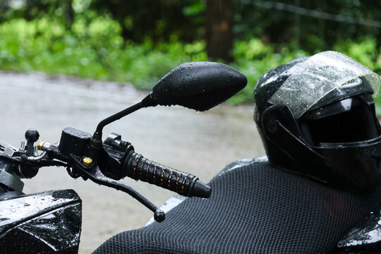 Rainy motorcycle scene with black helmet, mirror, handlebar, and textured seat beside wet road, creating dramatic outdoor travel mood