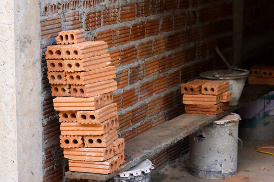 Stacked clay brick at unfinished construction site showing hollow masonry units, rough wall, cement bucket, and building materials