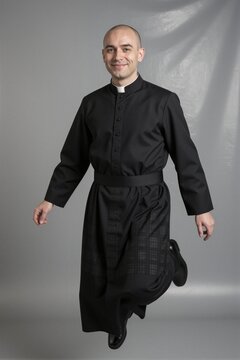 A man in a black clerical cassock poses mid-stride in a plain gray studio backdrop with soft lighting.