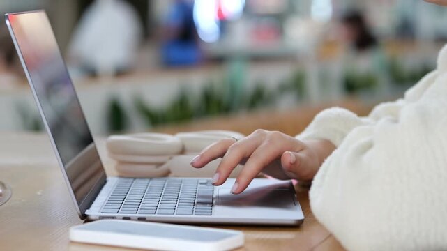 Person typing on laptop in cozy cafe setting. Fingers move across keyboard smoothly. Phone rests beside laptop on wooden table. Warm lighting surrounds workspace