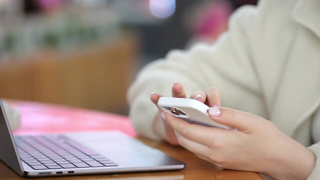 Close-up of person using smartphone and laptop on wooden table. Hands type on phone while laptop rests nearby. Soft background with warm lighting. Focus on multitasking with digital devices