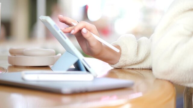 Person using tablet with stylus at wooden table. Warm lighting highlights cozy indoor setting. Soft focus on background objects including coffee cup