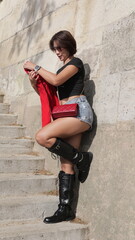 Cheerful Asian woman woman in a black t-shirt, denim shorts and black boots stands on a staircase leaning against a stone wall © iFocus