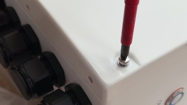 Close up of technician using insulated screwdriver to unscrew screw bolt from white electrical enclosure box cover. Hand unscrewing fastener on junction box, equipment maintenance concept