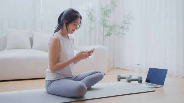 Asian woman resting after home workout and using mobile phone. Young sporty female after practicing yoga, break in doing exercise, relaxing on yoga mat, texting on the cell phone, holding smartphone