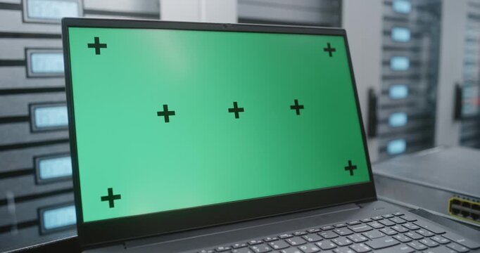Laptop With Green Screen Display Rests on Table Beside Server Racks in Data Center, Providing Clean Screen Replacement Footage for Software Interfaces, Dashboards and Enterprise Technology Visuals.